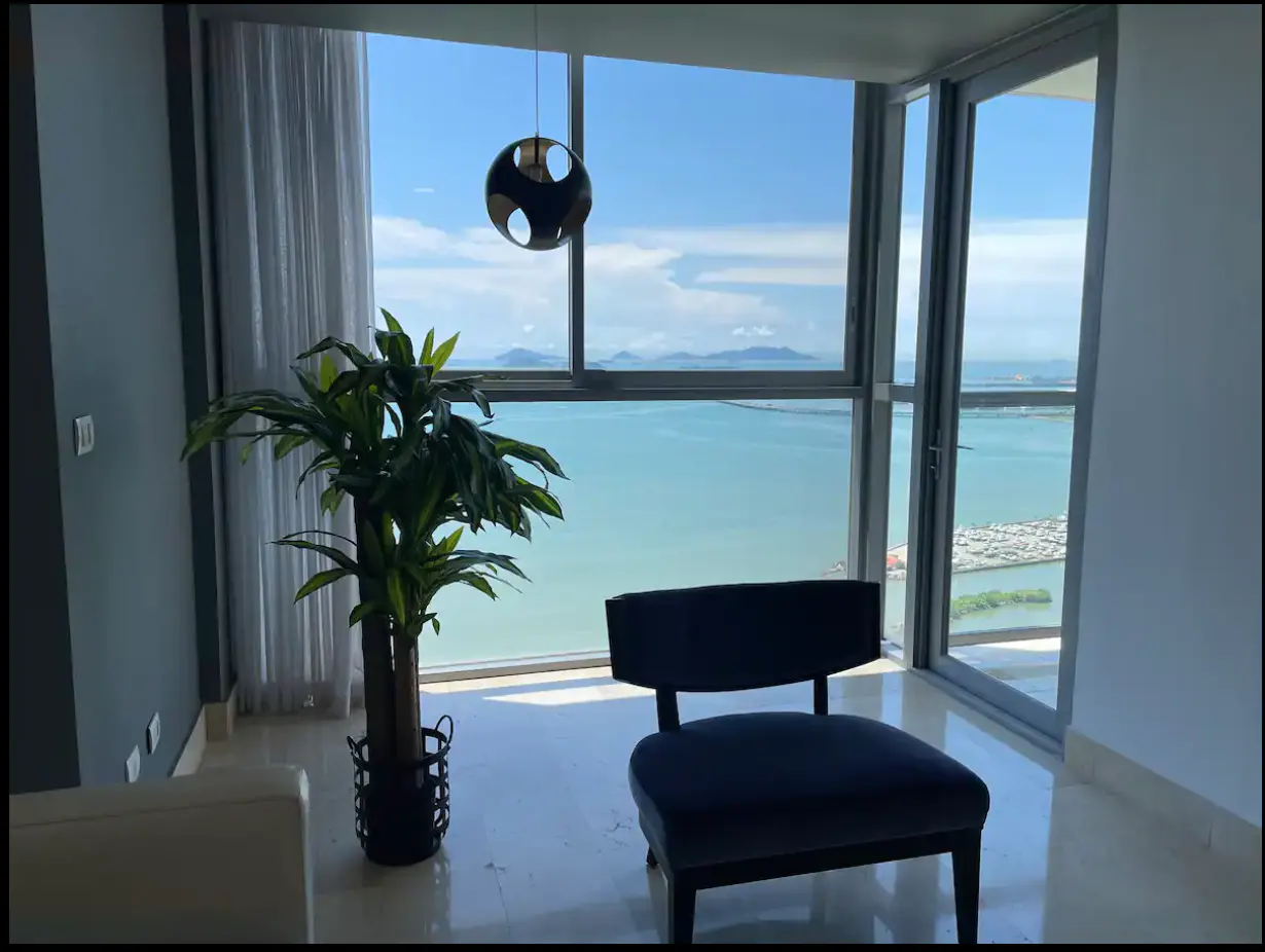 Bright room with ocean view. A black chair sits near a large window. A potted plant and modern light fixture add to the decor.