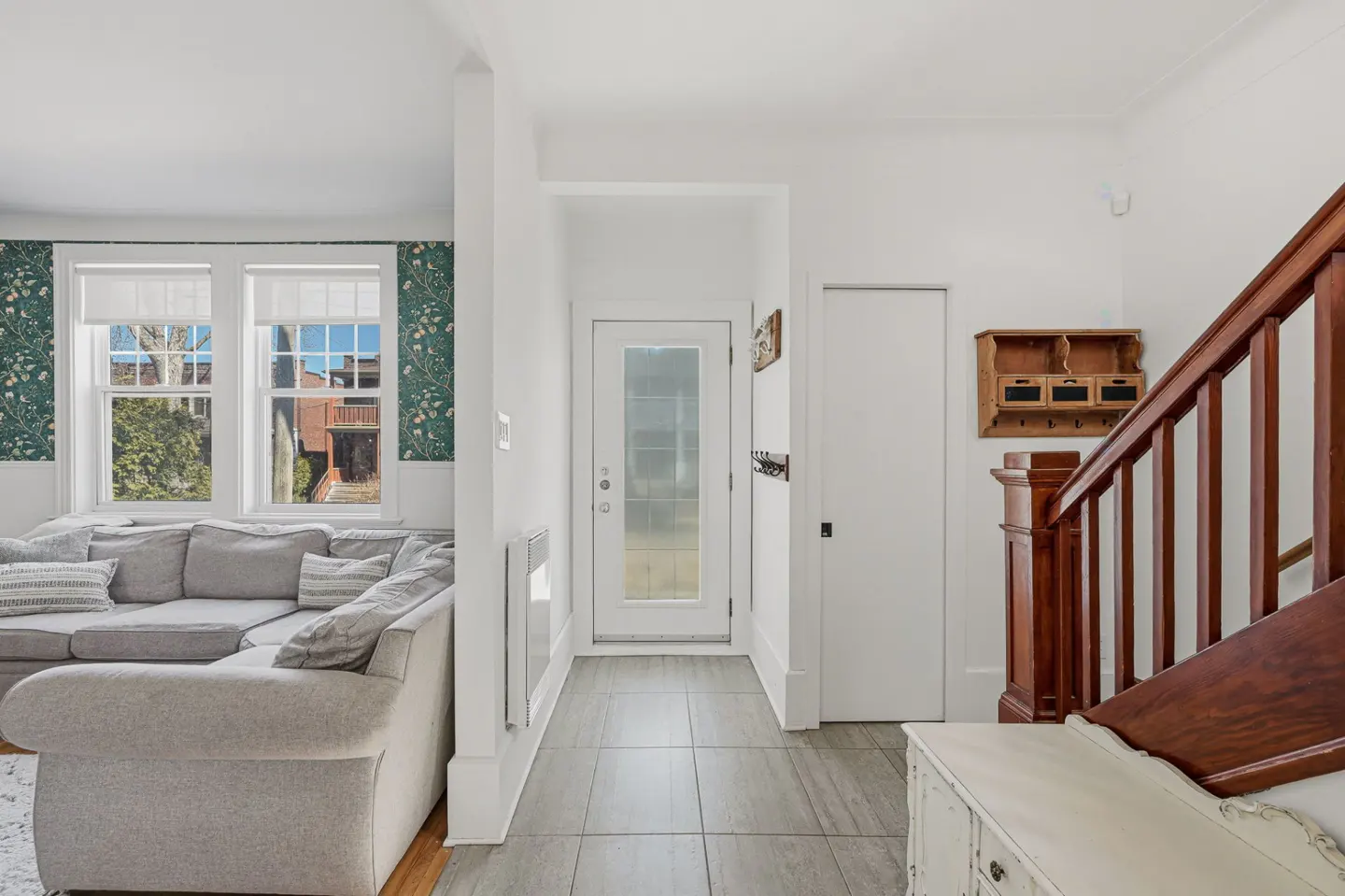 A bright, modern home interior with a gray sectional sofa, white walls, and a wooden staircase. A glass door leads to the outside.