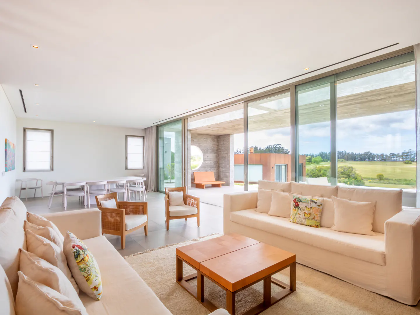 Bright, modern living room with white sofas, wood chairs, and a square coffee table. Large windows offer a view of a green landscape.