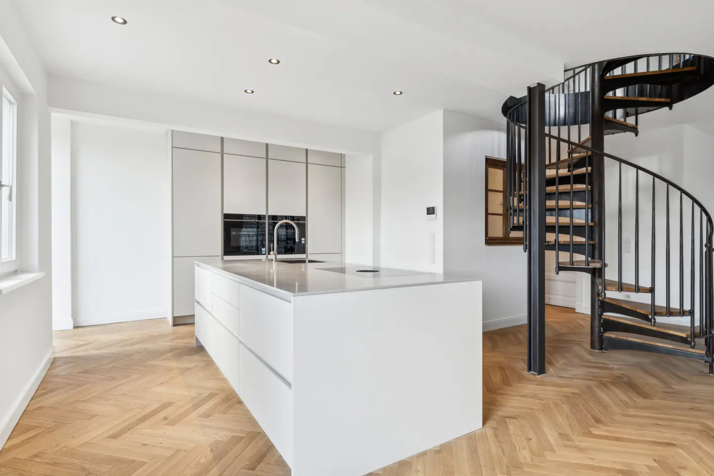 Bright, modern kitchen with white cabinets, island, and herringbone wood floors. A black spiral staircase is visible in the background.