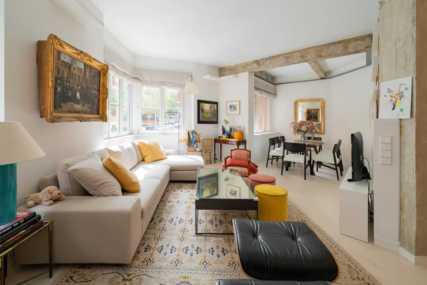 Bright living room with white walls, a beige sectional sofa with yellow pillows, and a patterned rug. A dining table and chairs are visible in the background.