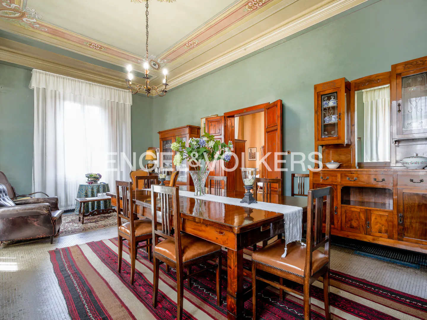 A dining room with a long wooden table, chairs, and a striped rug. A chandelier hangs above, and wooden cabinets line the walls.