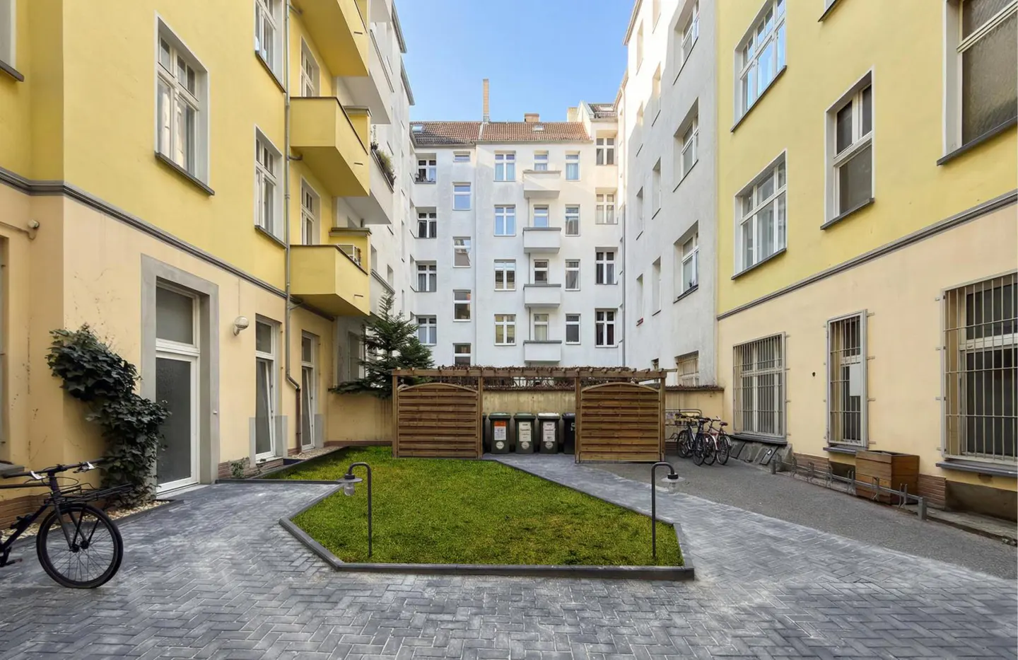 Apartment courtyard with yellow and white buildings, gray brick path, and small green lawn. Bicycles are parked in the background.