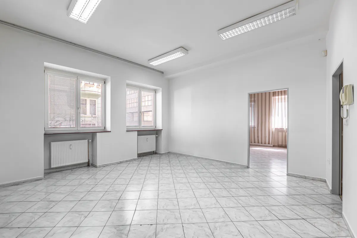 Bright, empty office space with white walls, tiled floor, and three windows. A doorway leads to another room with striped curtains.