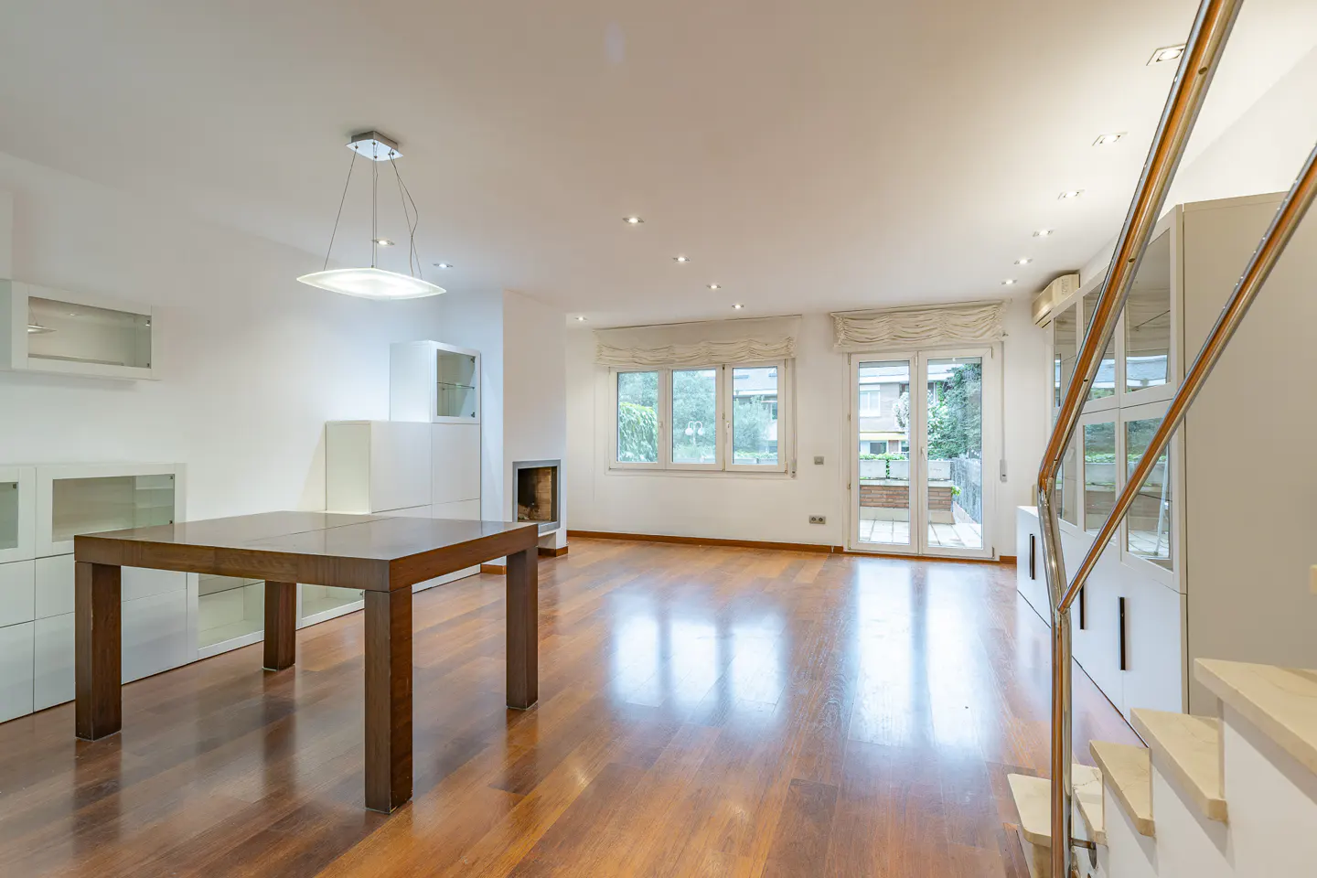 Bright, modern living room with hardwood floors, a dark wood table, white walls, and a staircase with metal railing.
