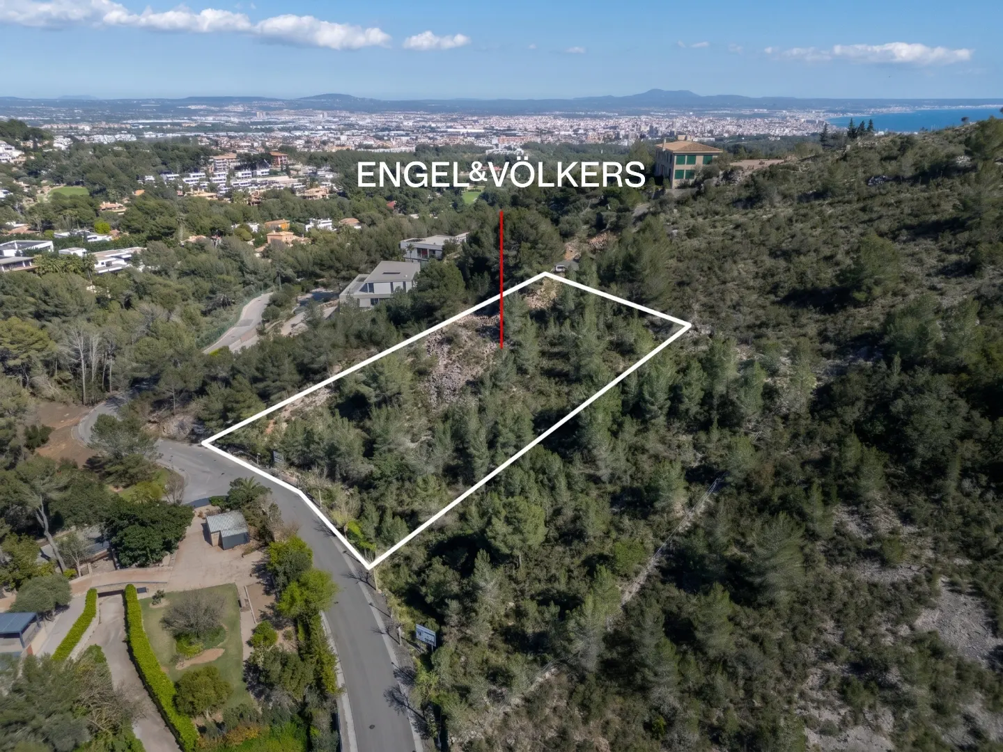 Aerial view of a wooded lot outlined in white, with Engel & Völkers logo, on a hillside overlooking a city and the sea.
