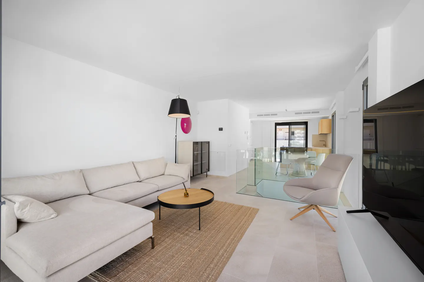 Bright, modern living room with a beige sectional sofa, round wood coffee table, and a tan rug. A glass railing separates the living area from the dining space.