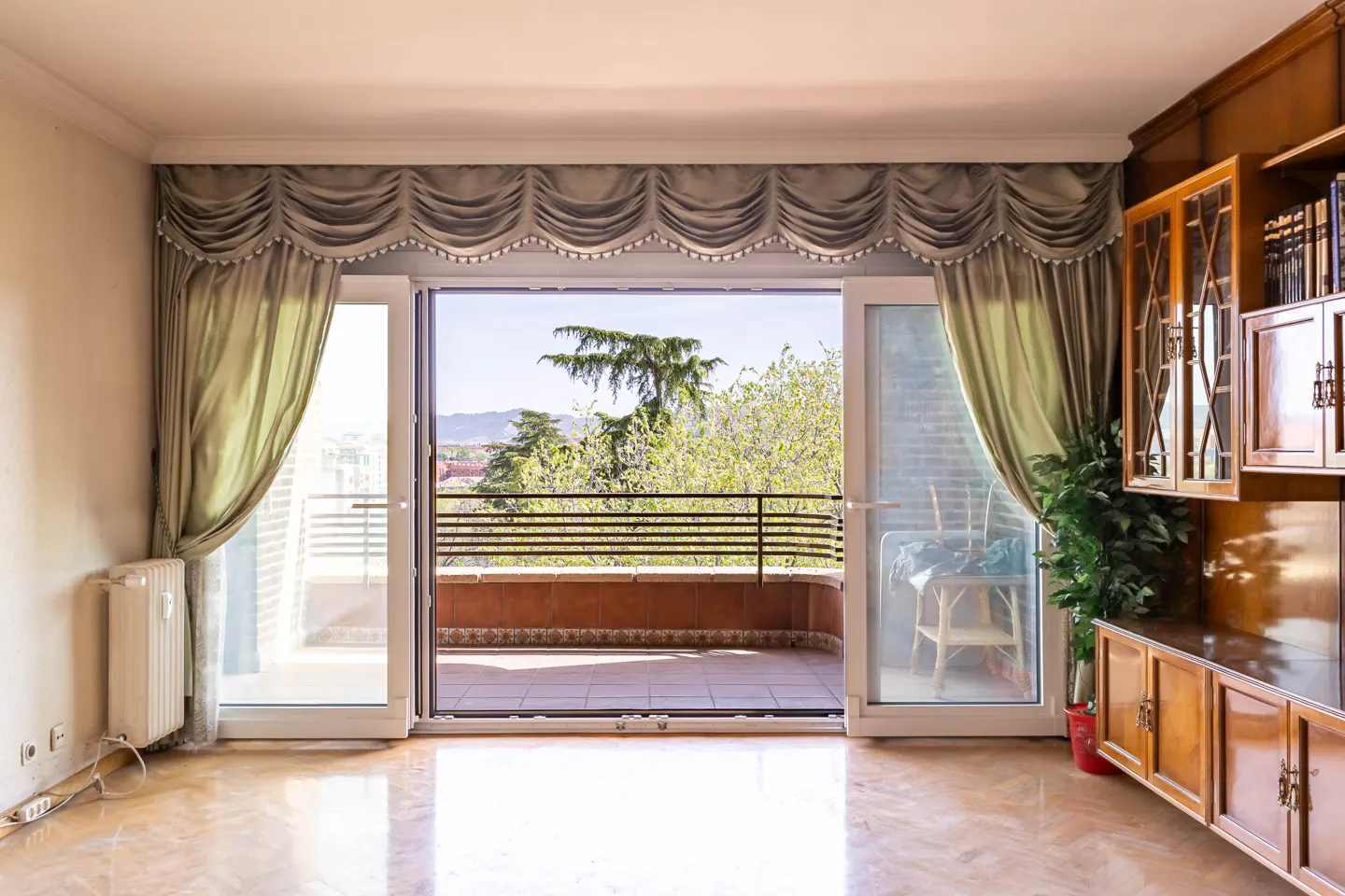 Living room with open sliding doors to a balcony with trees and a mountain view. Draped curtains frame the doors.