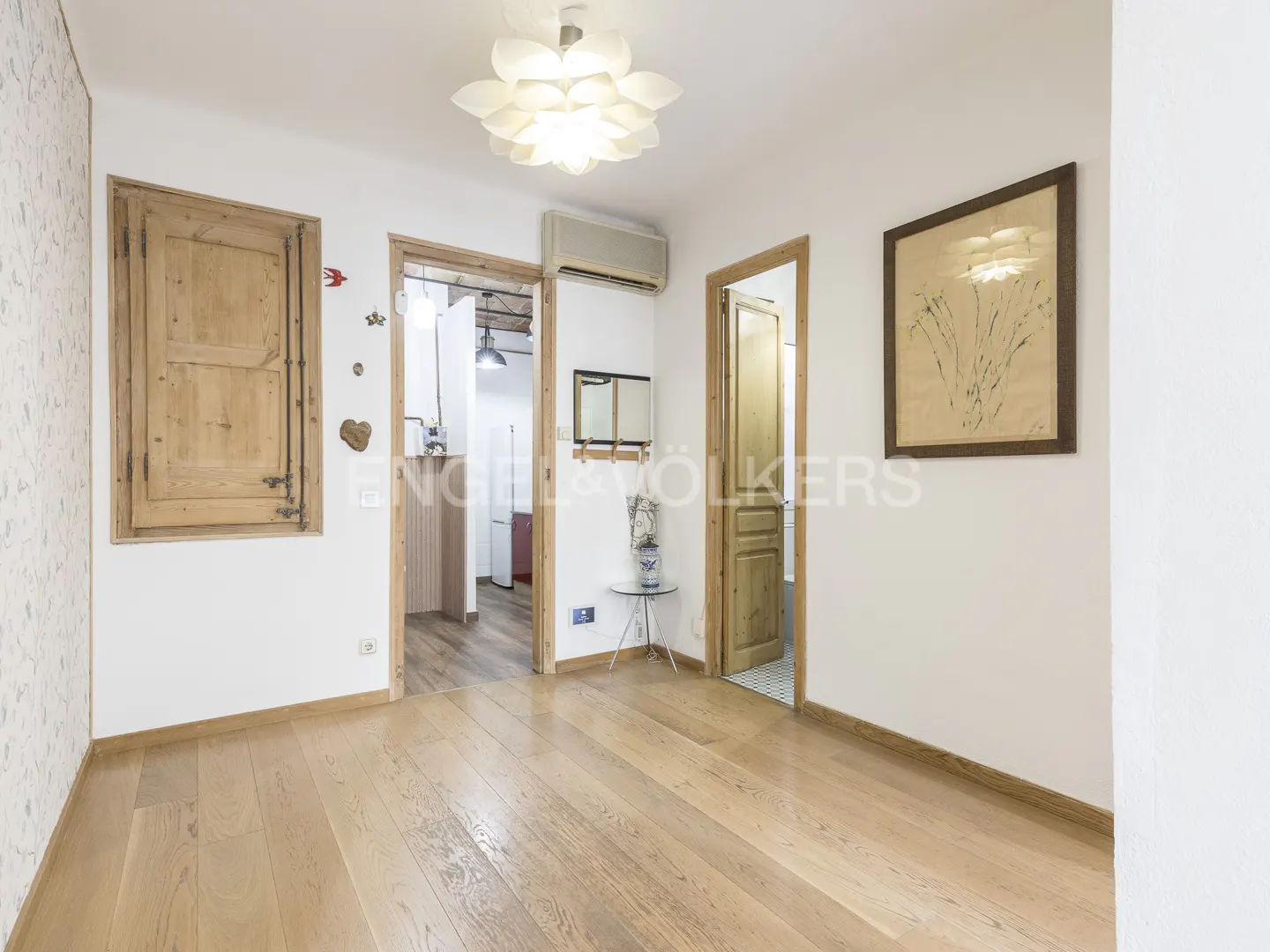 Bright apartment interior with light wood floors, white walls, and wooden doors. A floral light fixture hangs from the ceiling.