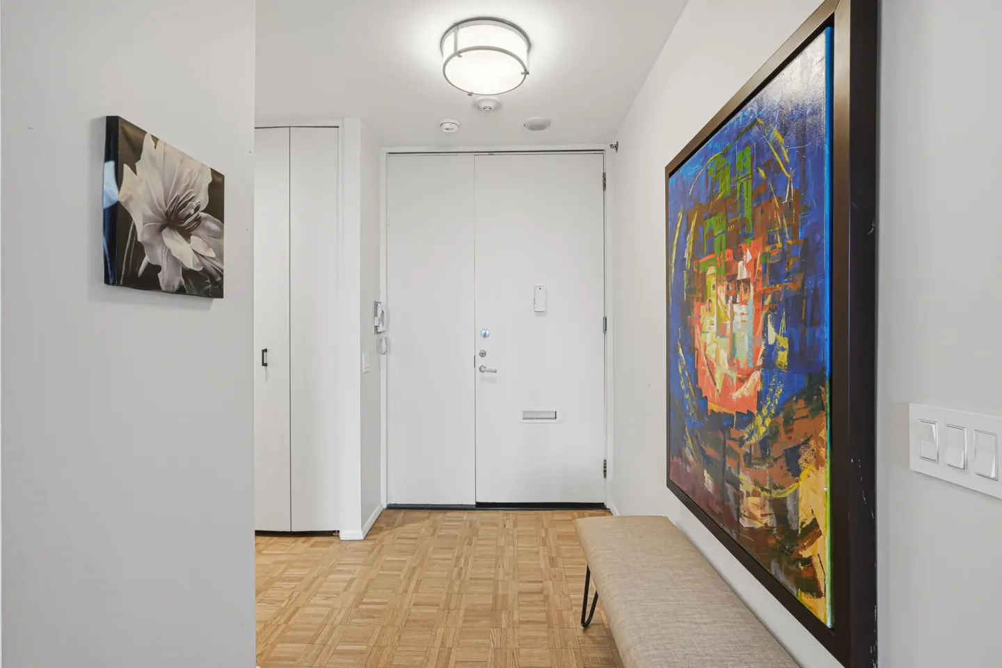 Hallway with parquet floors, white walls, and a white front door. A colorful painting hangs above a bench. A black and white flower print is on the left wall.