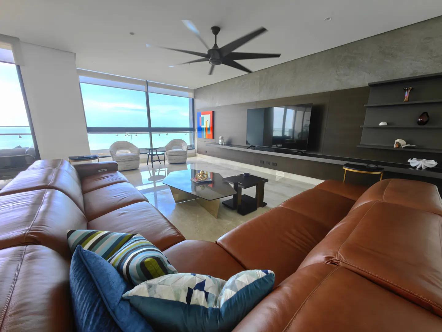 A modern living room with a brown leather sectional sofa, blue pillows, and a view of the ocean through large windows.