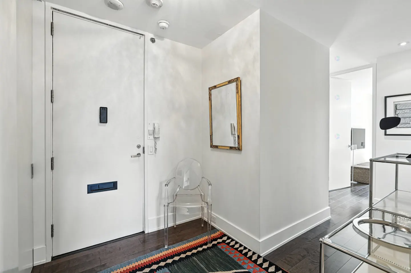 Entryway with white door, dark wood floors, and white walls. A clear chair sits near a gold-framed mirror above a colorful rug.