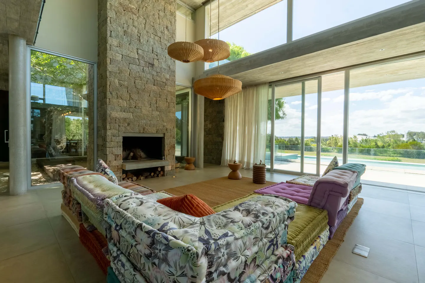 Bright living room with stone fireplace, floor-to-ceiling windows, and a colorful sectional sofa. Three woven pendant lights hang from the concrete ceiling.