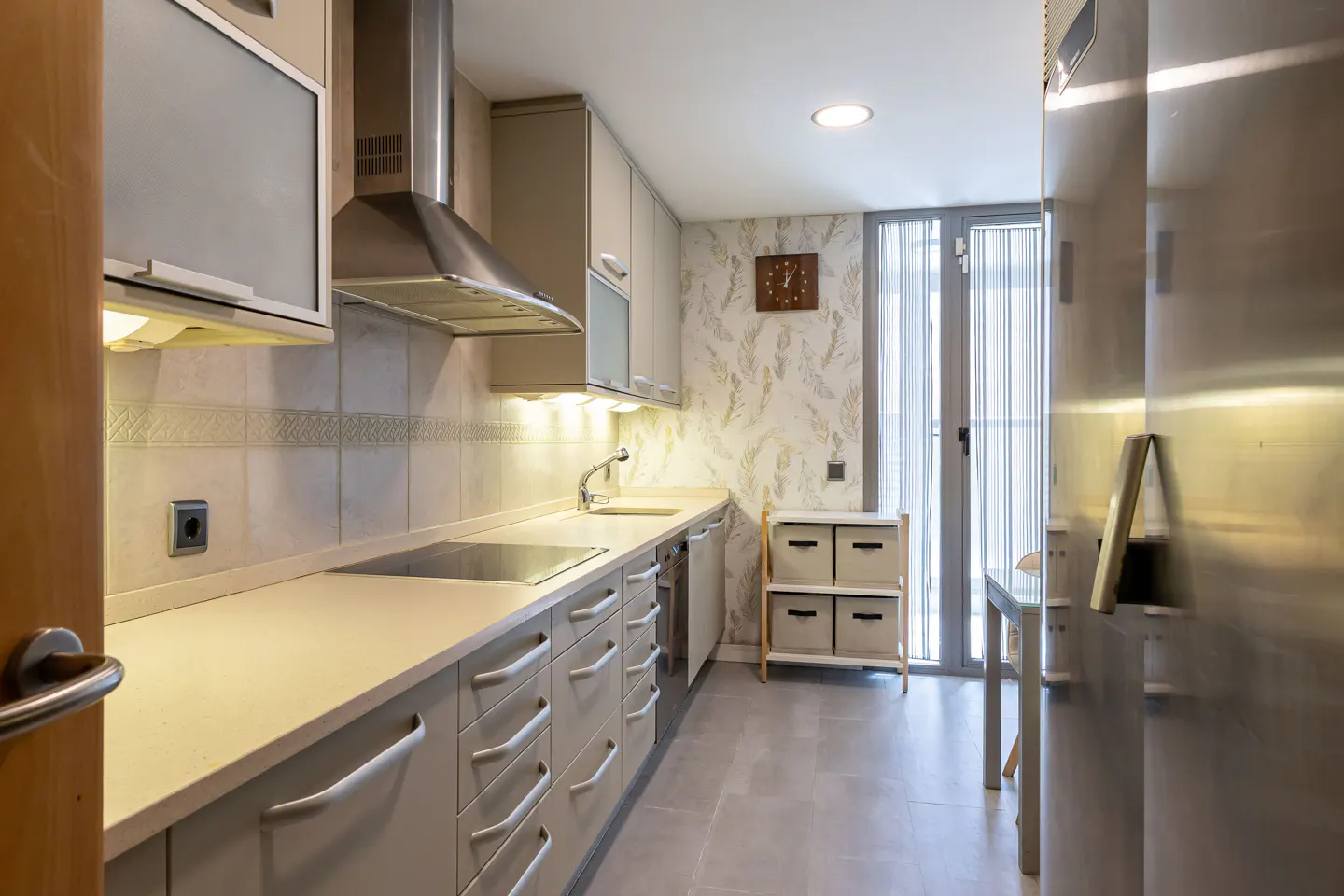 A modern kitchen with gray cabinets, stainless steel appliances, and light countertops. A storage unit sits near a glass door with vertical blinds.