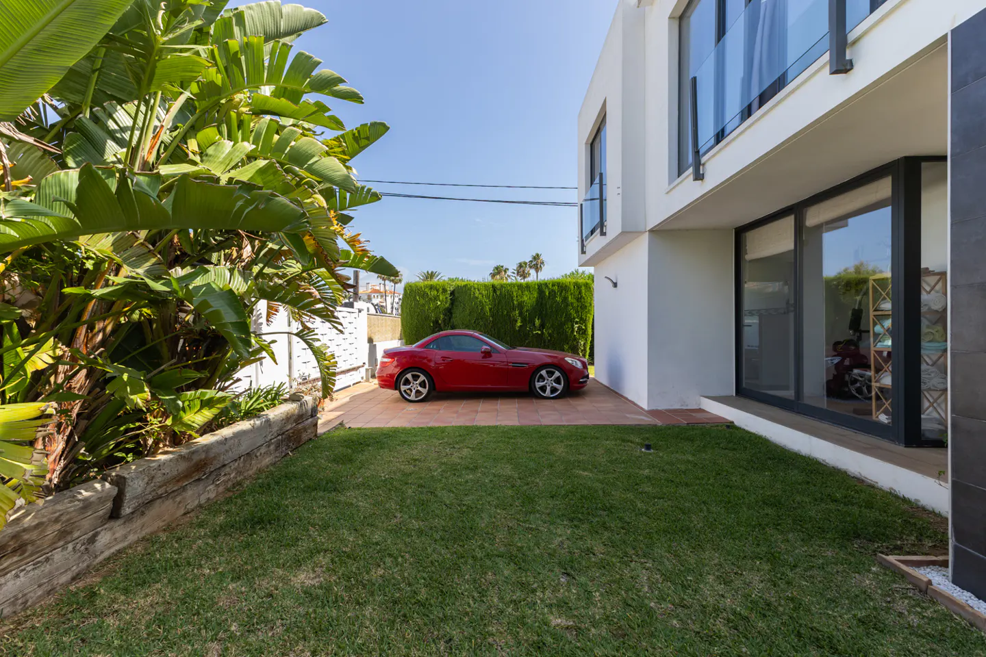 A red convertible parked on a tiled driveway next to a modern white house with a green lawn and large tropical plants.