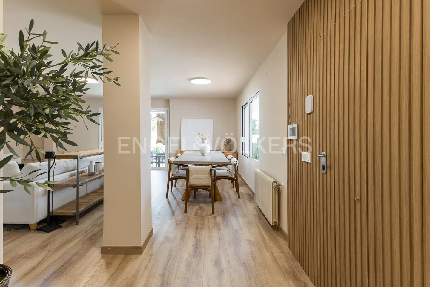A bright, modern home interior with a dining table and chairs visible down a hallway. A wood-paneled door is on the right.