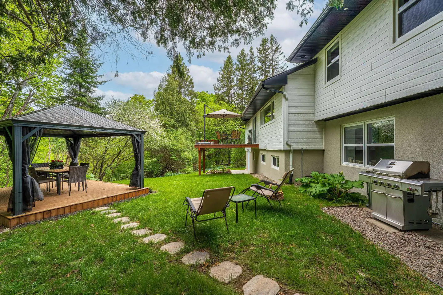 Backyard with a gazebo, stone path, lawn chairs, a stainless steel grill, and a raised deck against a white house.