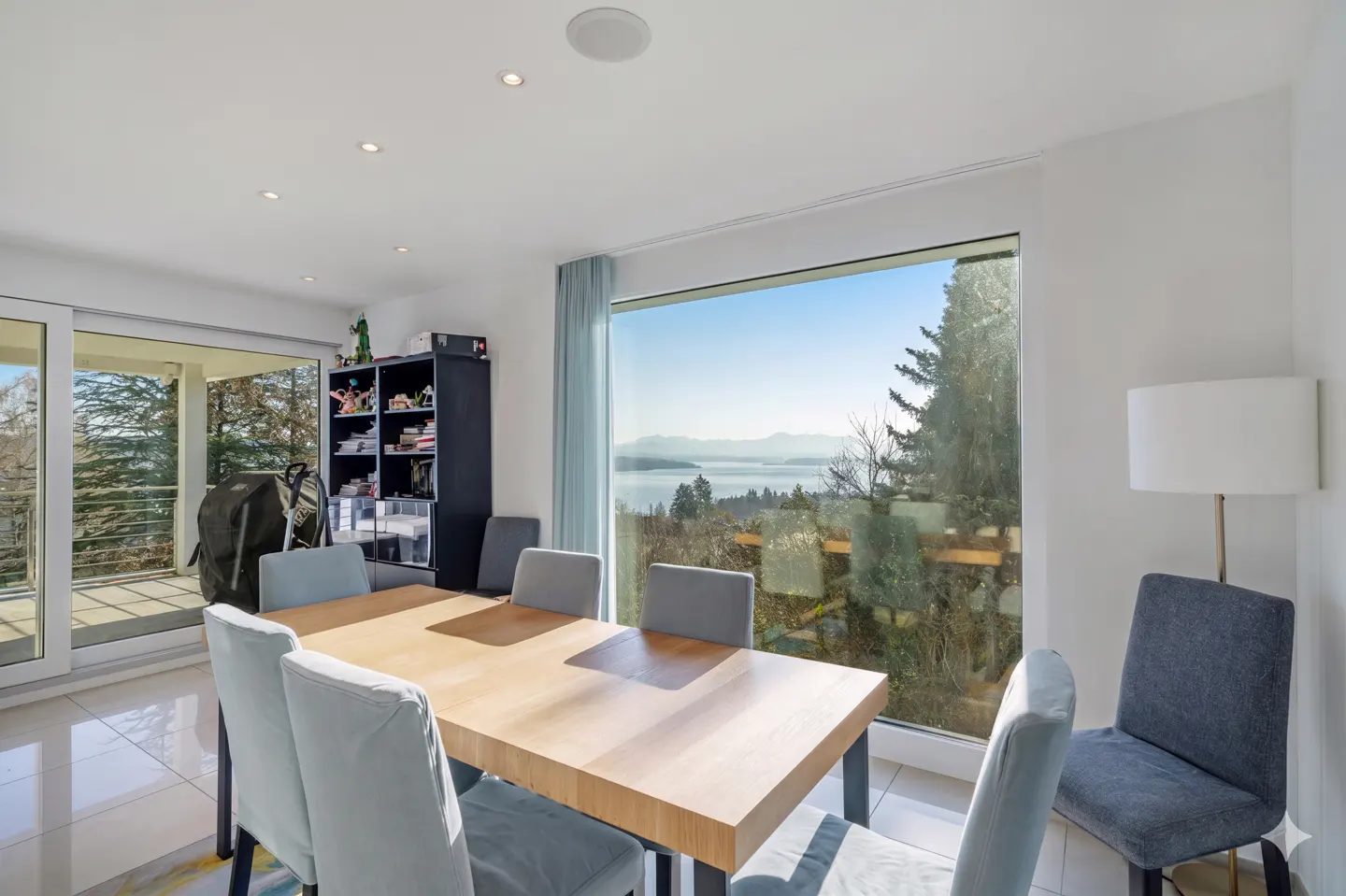 Bright dining room with a wood table, blue chairs, and a large window with a scenic view of a lake and mountains.