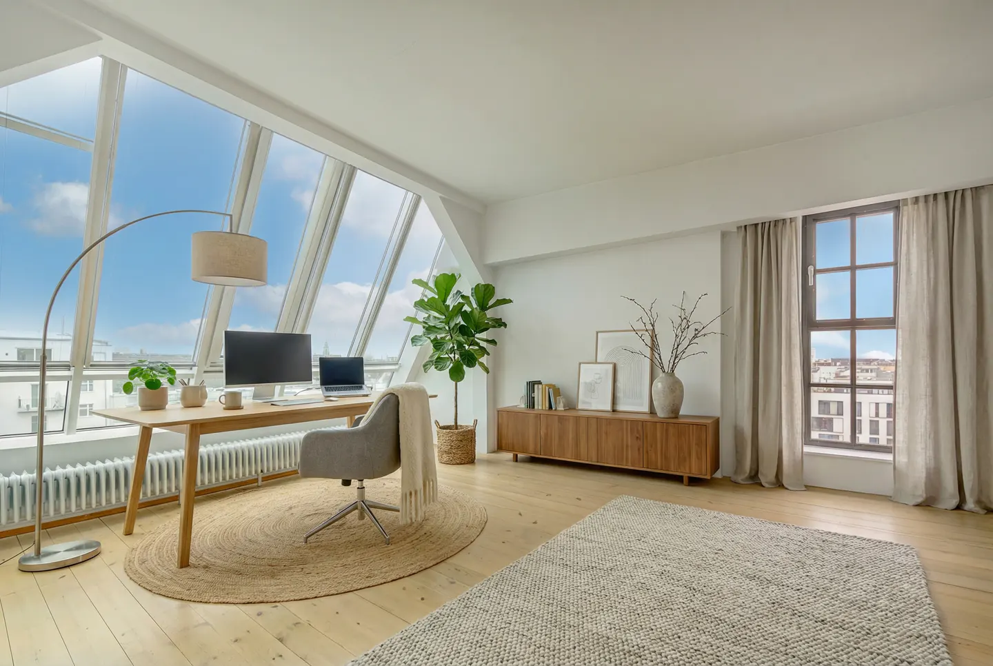 Bright home office with a wooden desk, grey chair, large windows, a fiddle leaf fig plant, and city views.