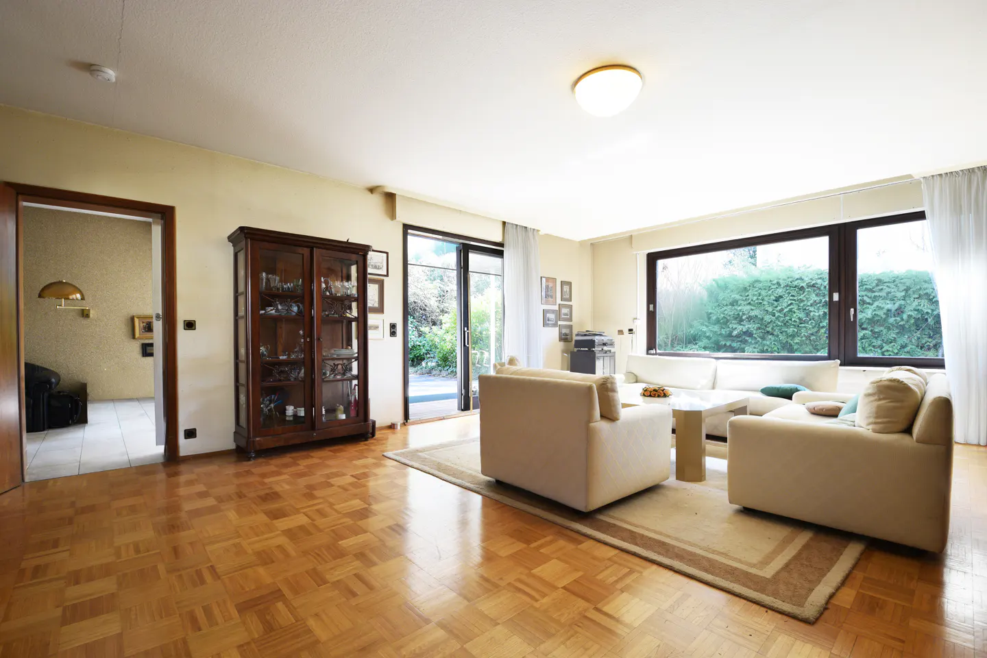 A bright living room with parquet floors, a glass cabinet, and a white sofa set near a large window.