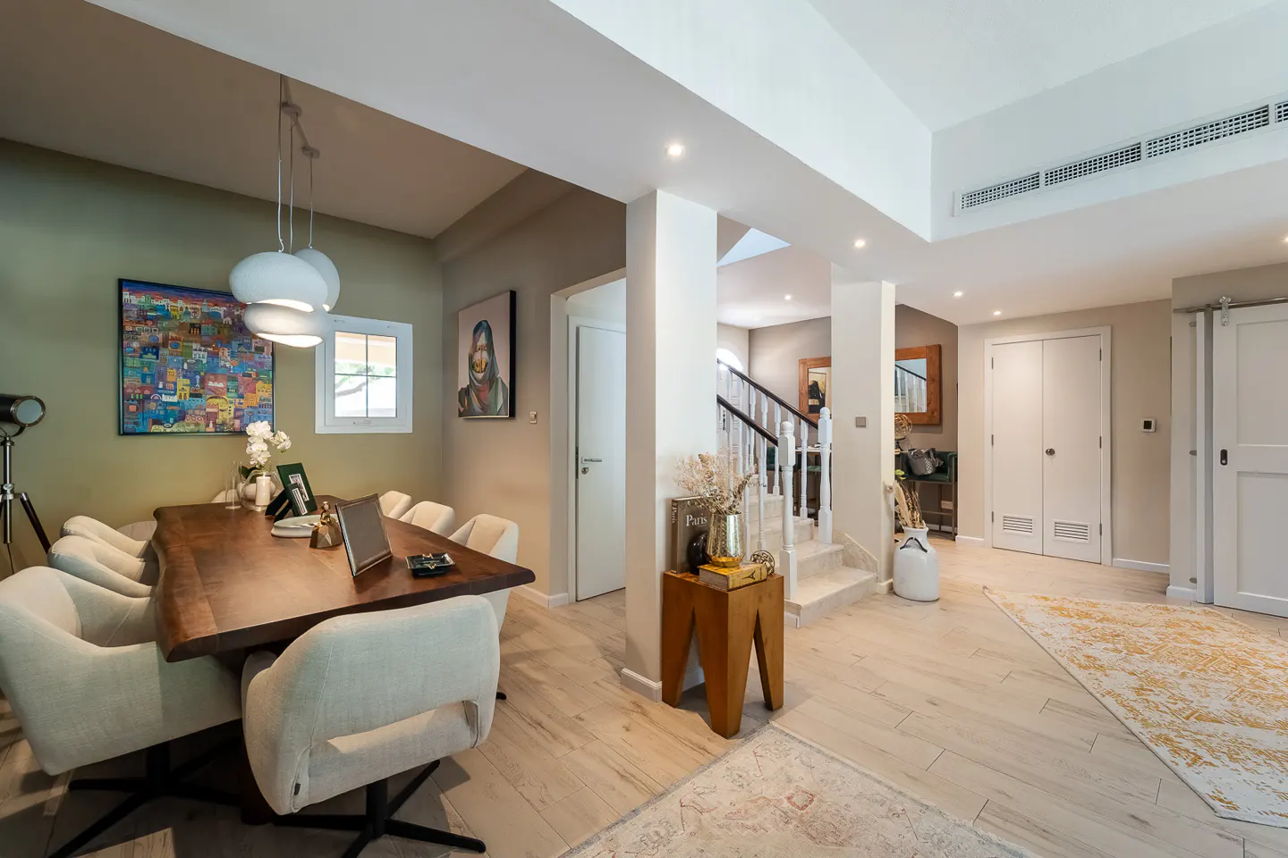 A bright, modern dining room with a wood table, white chairs, and artwork on the walls. A staircase leads to the upper level.