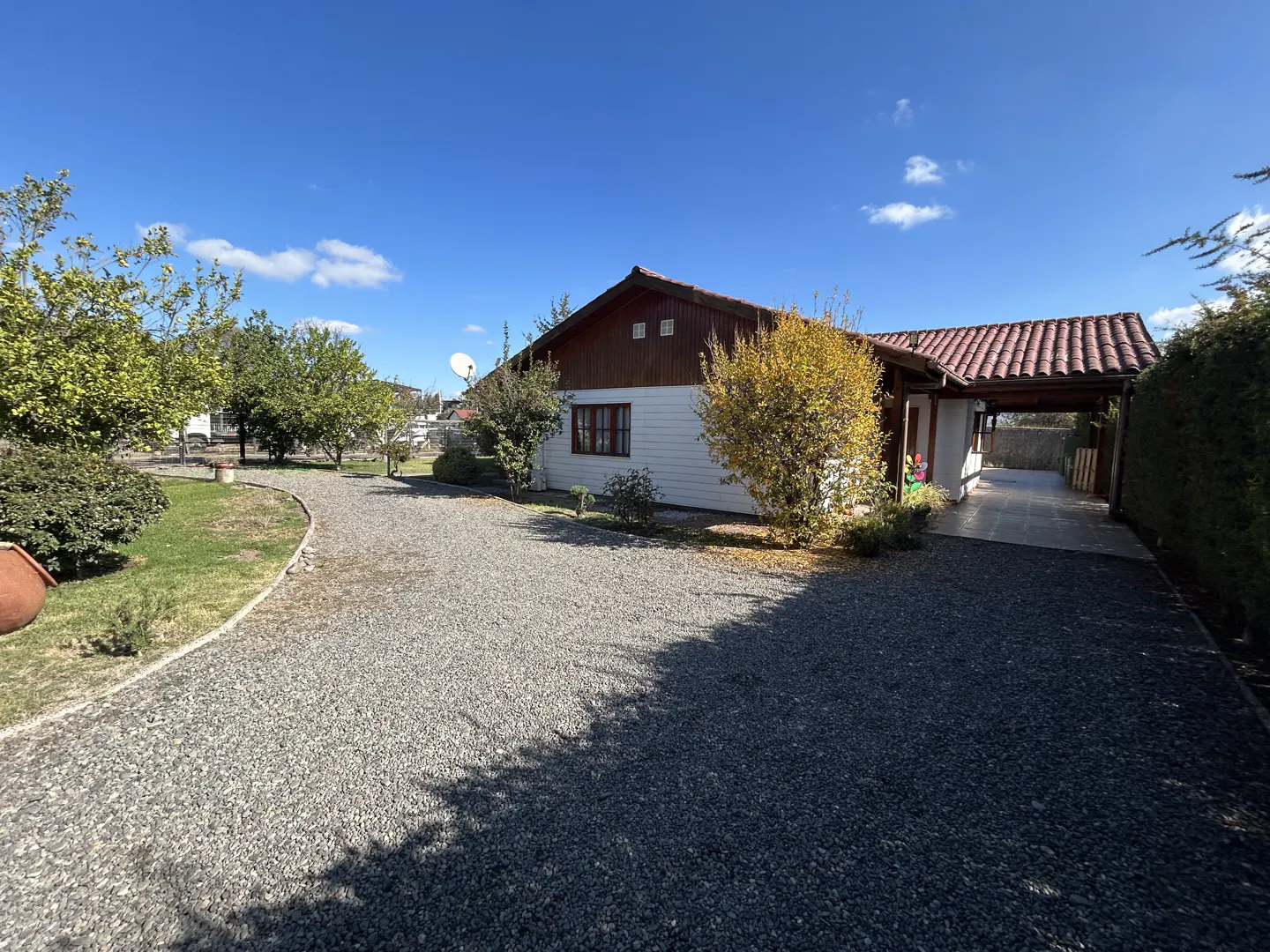 A single-story white house with a brown roof and trim, a gravel driveway, and green trees under a blue sky.