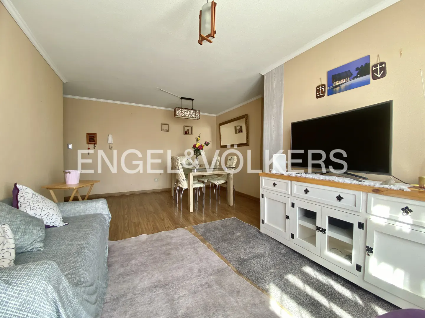 Living room with a gray sofa, dining table, and white TV stand with a large television on top. The walls are beige.