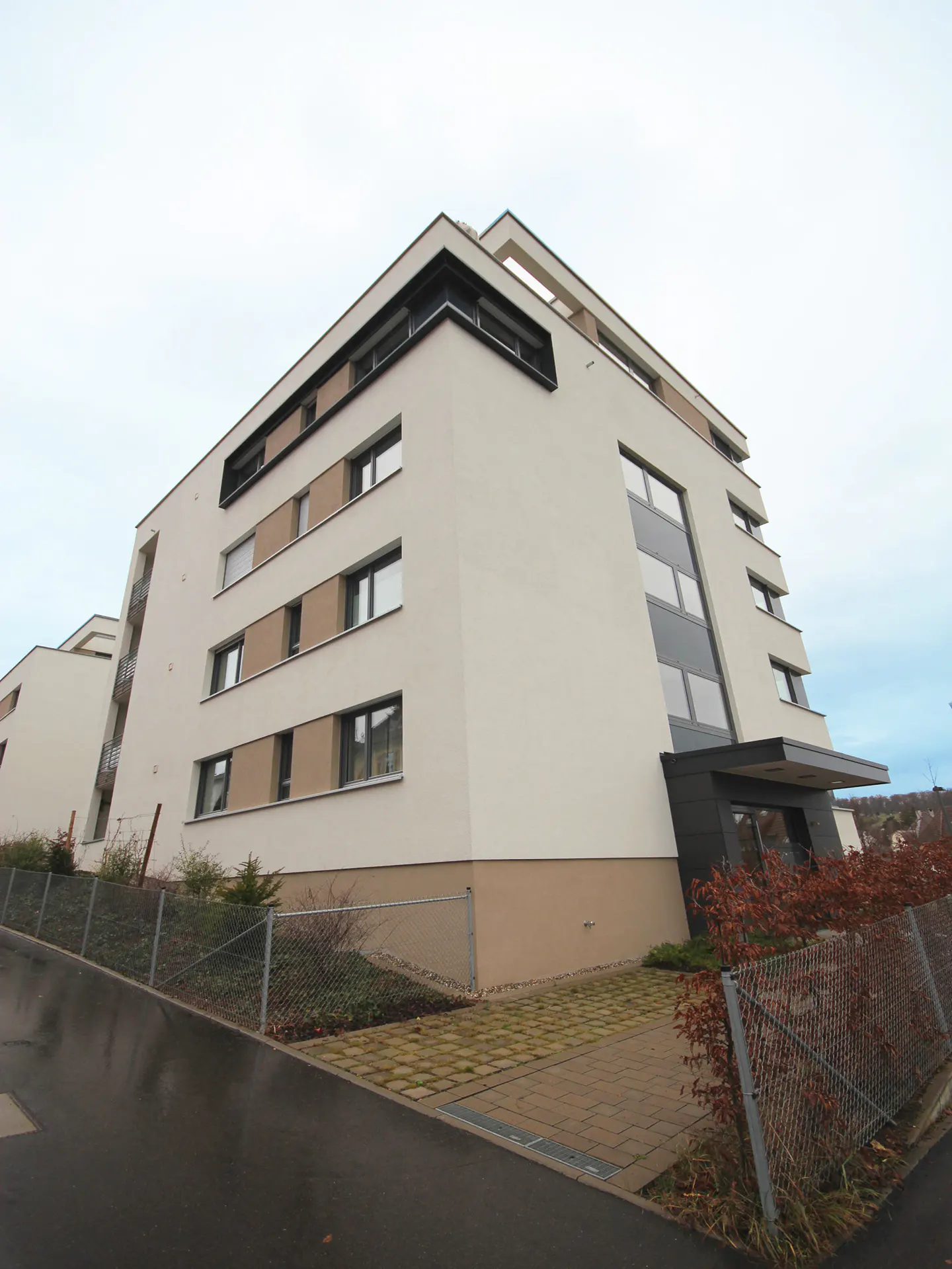 Modern apartment building with white and tan facade, black window frames, and a flat roof. A paved walkway leads to the entrance.