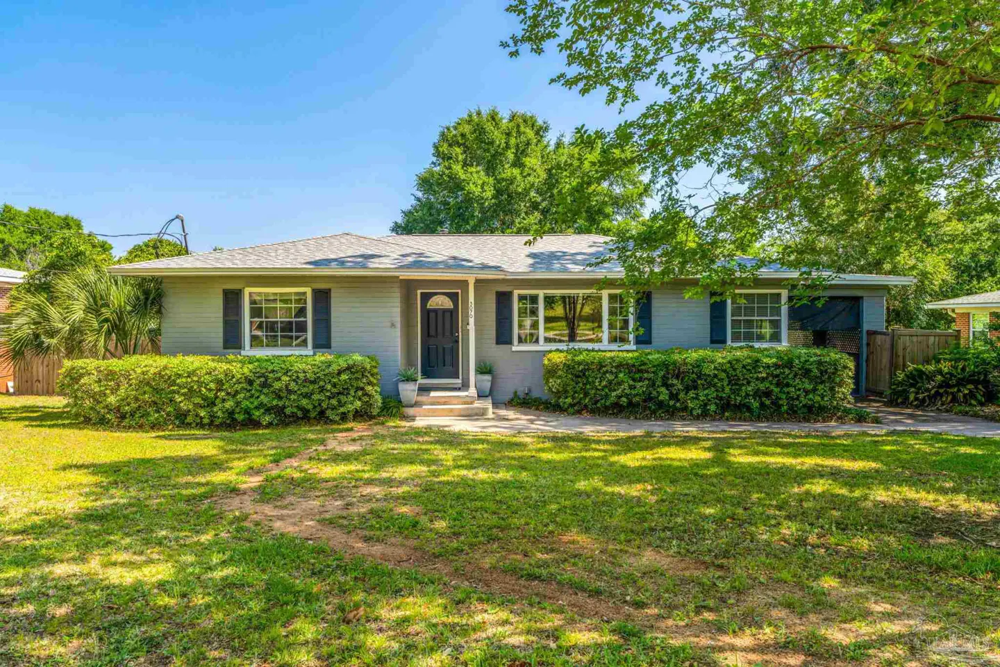 A single-story gray house with dark shutters and a black front door, surrounded by green bushes and a lawn.