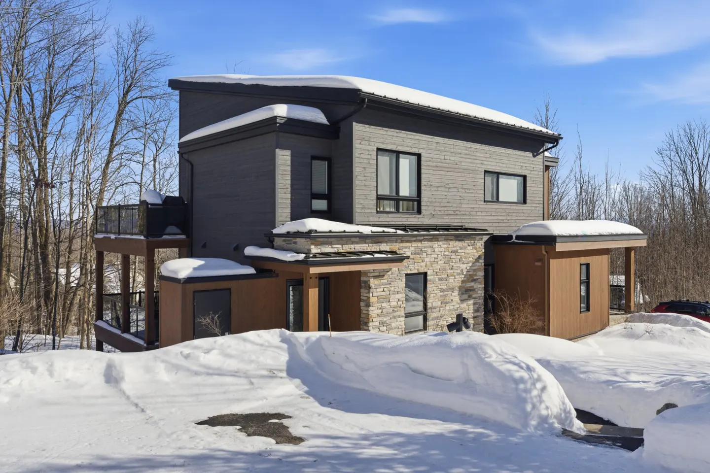 Modern two-story home with gray siding, stone accents, and snow-covered roof. Blue sky and bare trees in the background.