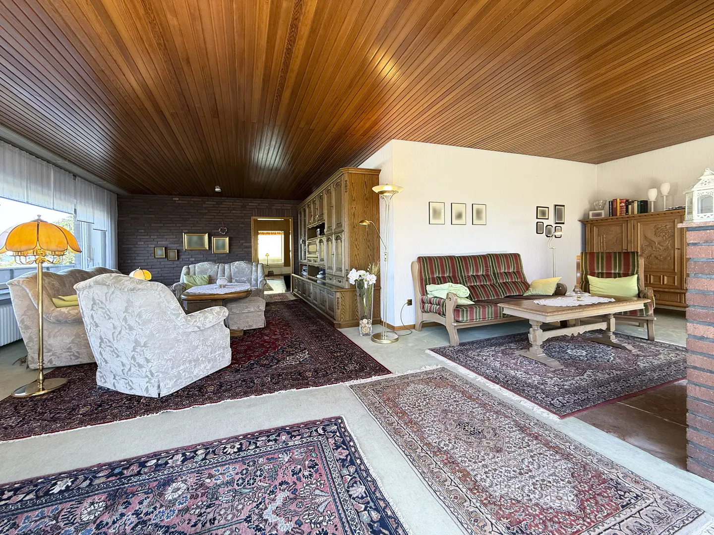Spacious living room with wood-paneled ceiling, patterned area rugs, vintage sofas, and a large wooden wall unit.