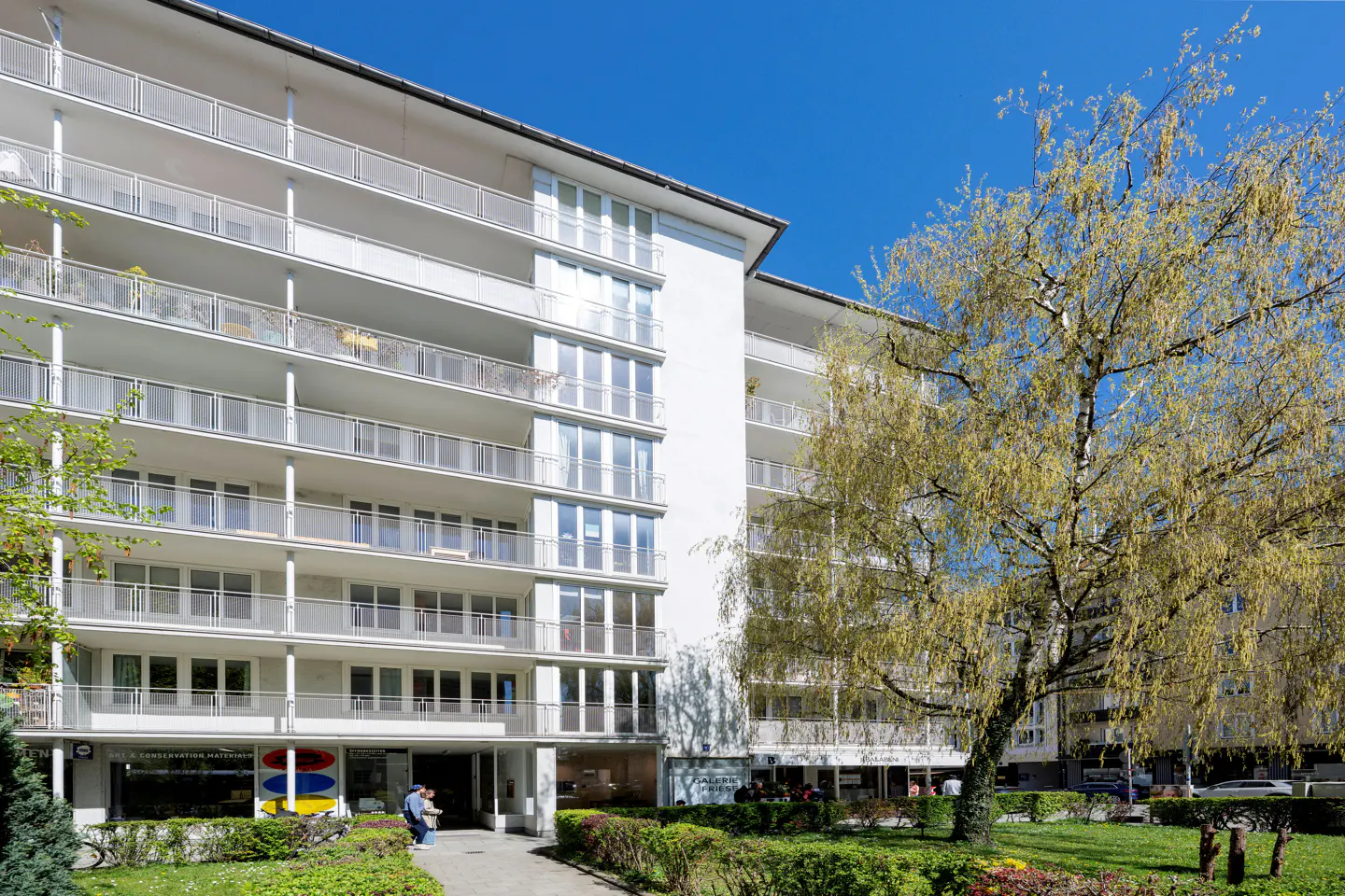 Exterior of a white, multi-story apartment building with balconies and a tree in front.