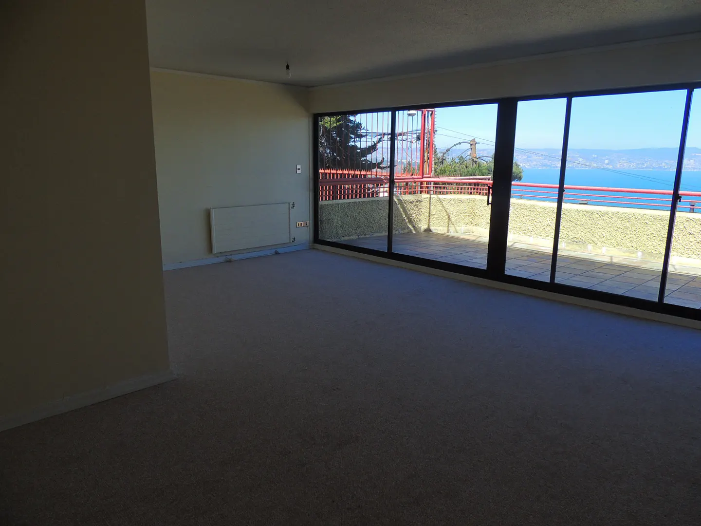 Empty room with beige carpet and walls. Large sliding glass doors open to a balcony with a red railing and a view of the ocean.