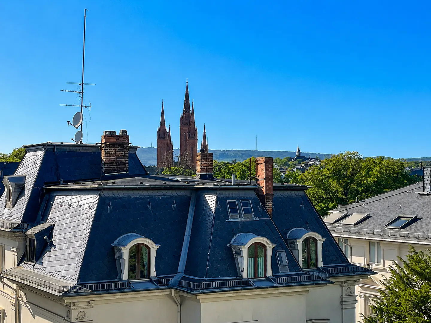 View of rooftops with dormer windows, slate roofs, and brick chimneys, with church spires in the background under a clear blue sky.