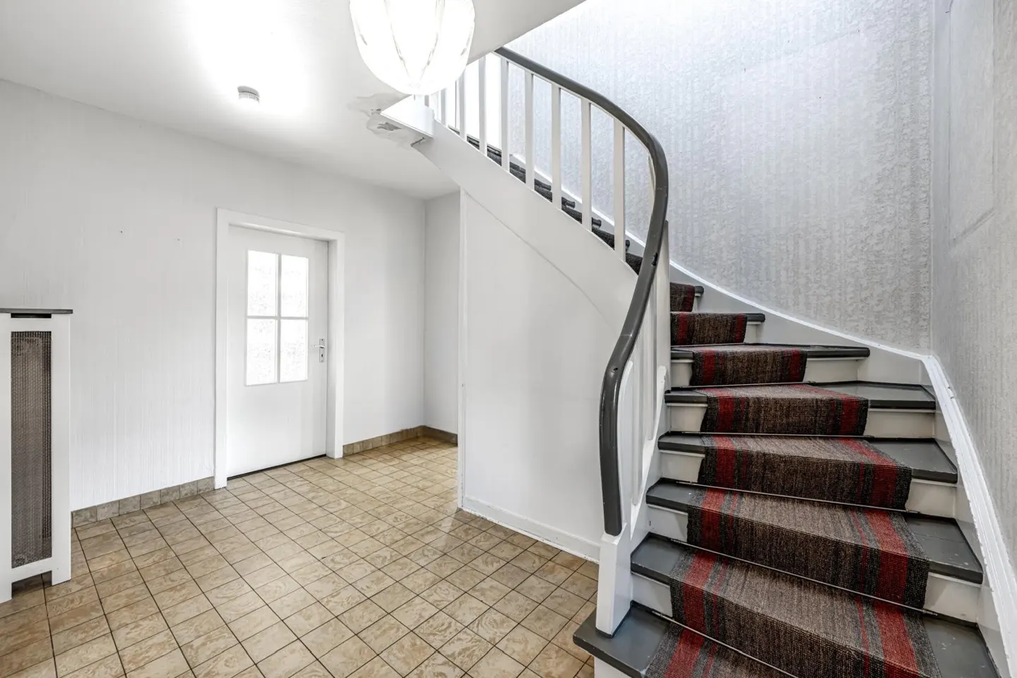 Bright foyer with a curved staircase. White walls, tiled floor, and a white door. The stairs have a red and brown carpet.