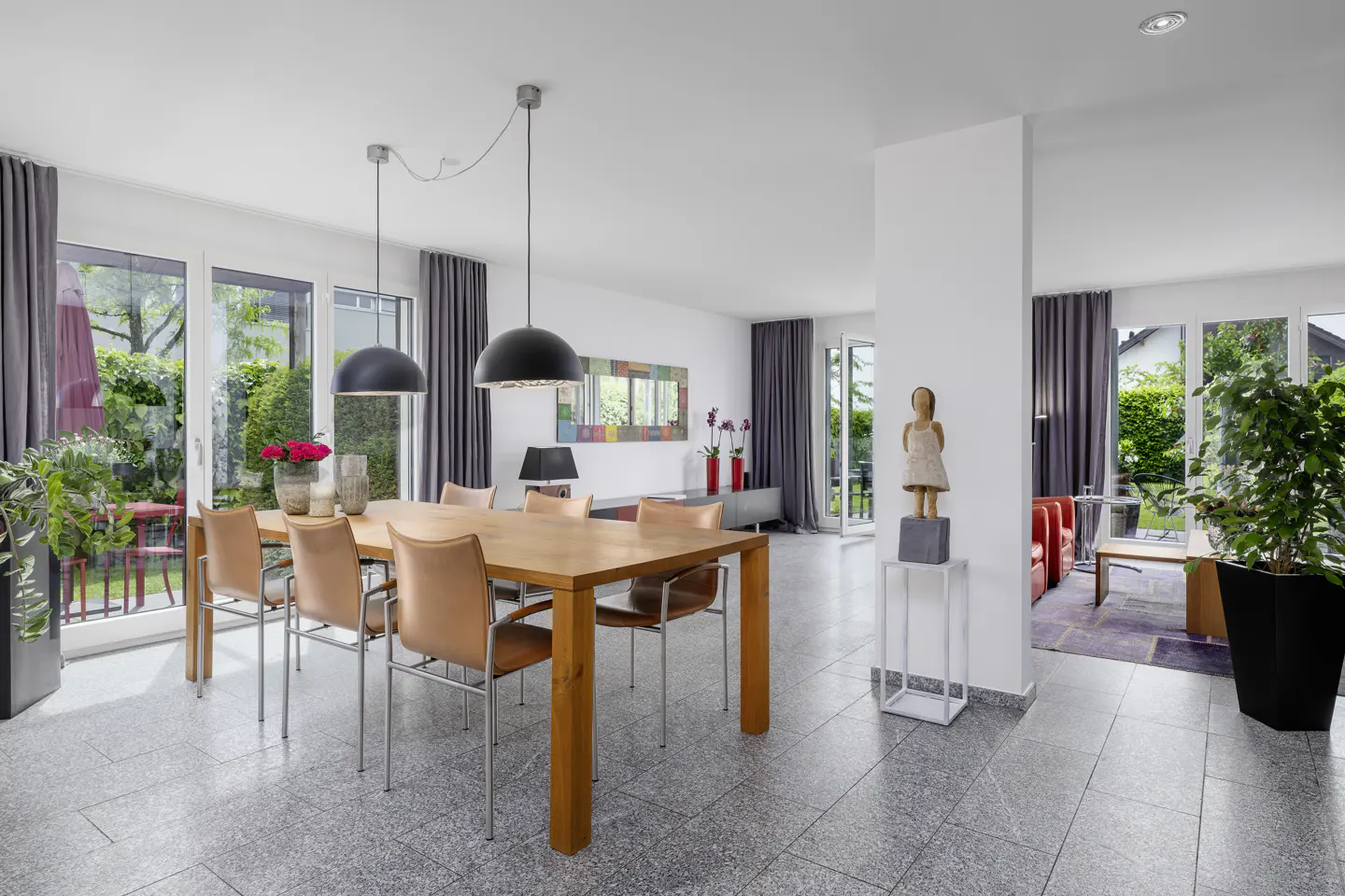 Bright, open dining room with a wood table, leather chairs, and black pendant lights. Gray curtains frame the windows overlooking a green garden.
