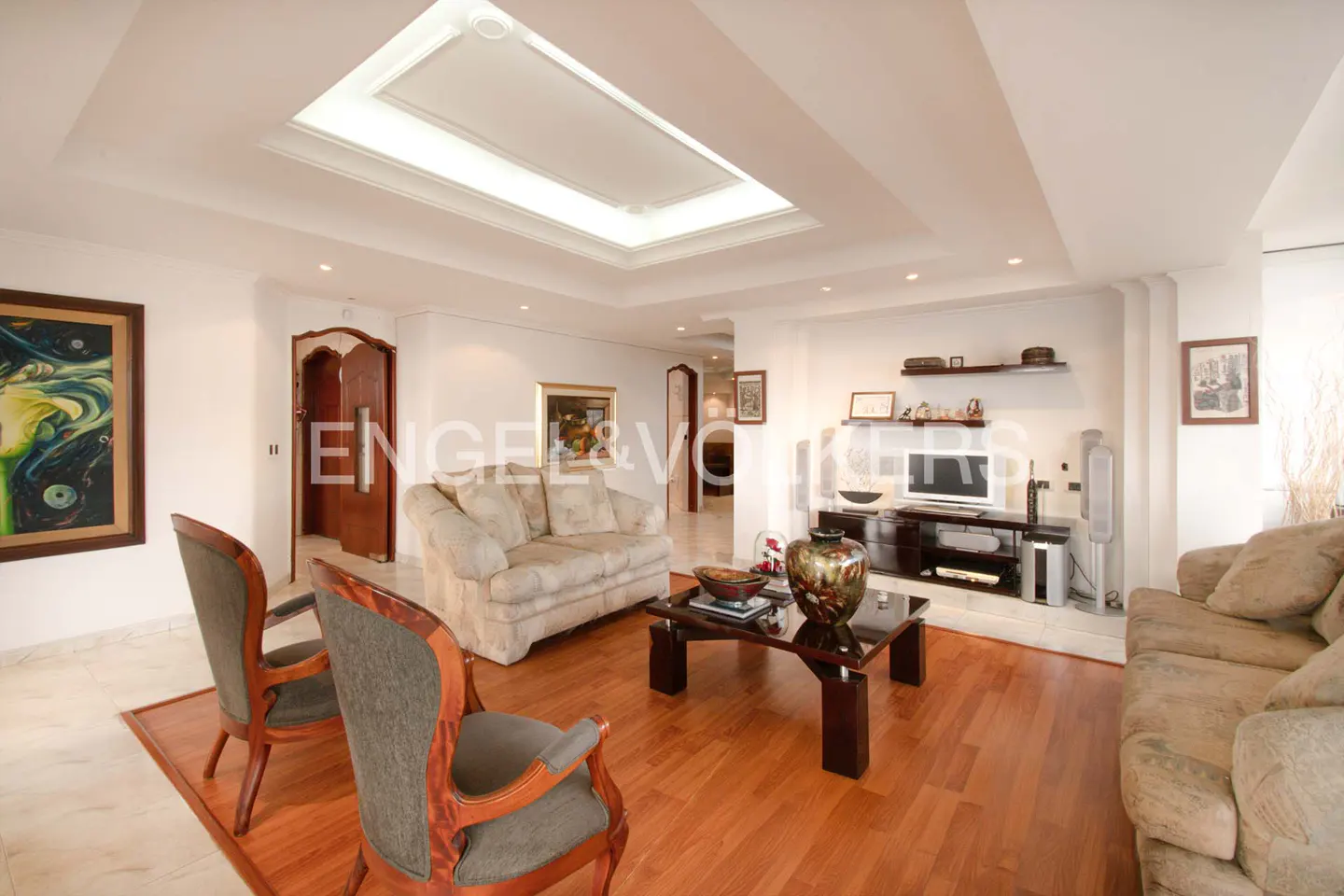 Living room with hardwood floors, beige sofas, two chairs, and a dark wood coffee table. A TV and shelves are against the wall.