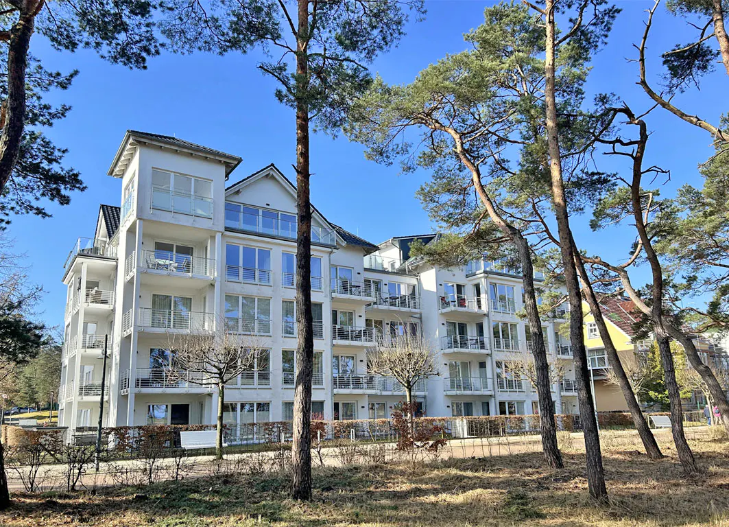 Modern white apartment building with balconies, framed by tall pine trees against a clear blue sky.