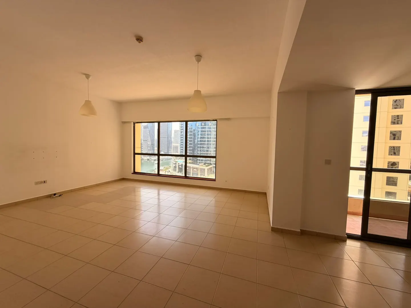 Empty room with beige tile floor, white walls, and two pendant lights. A large window shows a city skyline and a balcony door is on the right.