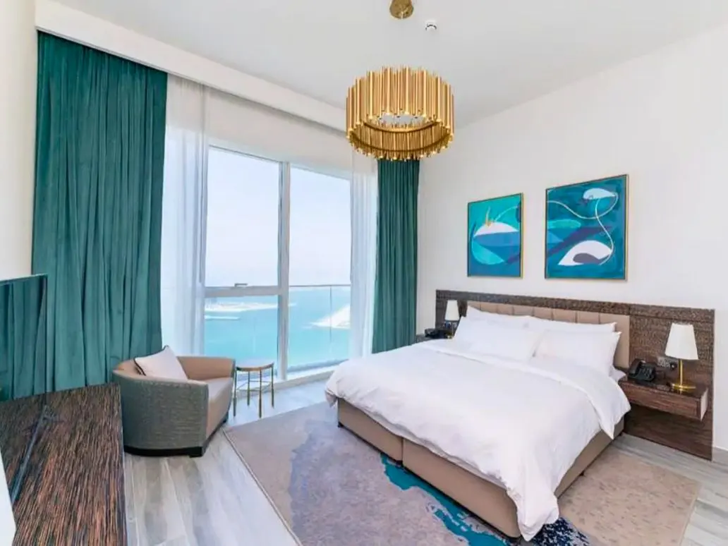Bright bedroom with ocean view. White linens on a king bed, teal curtains, and a gold chandelier. Two blue abstract paintings hang above the bed.