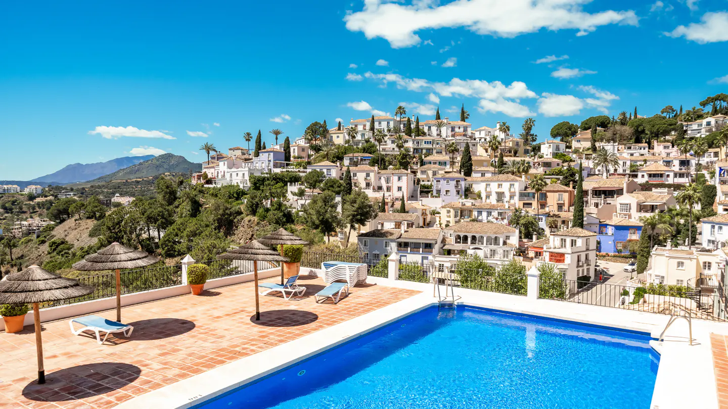 Rooftop pool with blue water, lounge chairs, and straw umbrellas. Hillside town and mountains in the background under a blue sky.