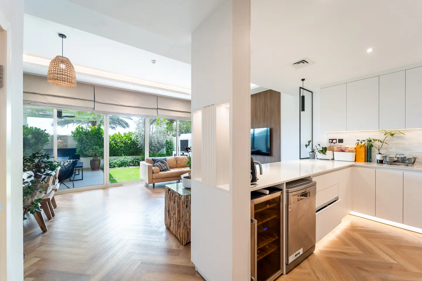 Open-concept living space with a white kitchen, wood floors, and a tan leather sofa visible through sliding glass doors to a green lawn.
