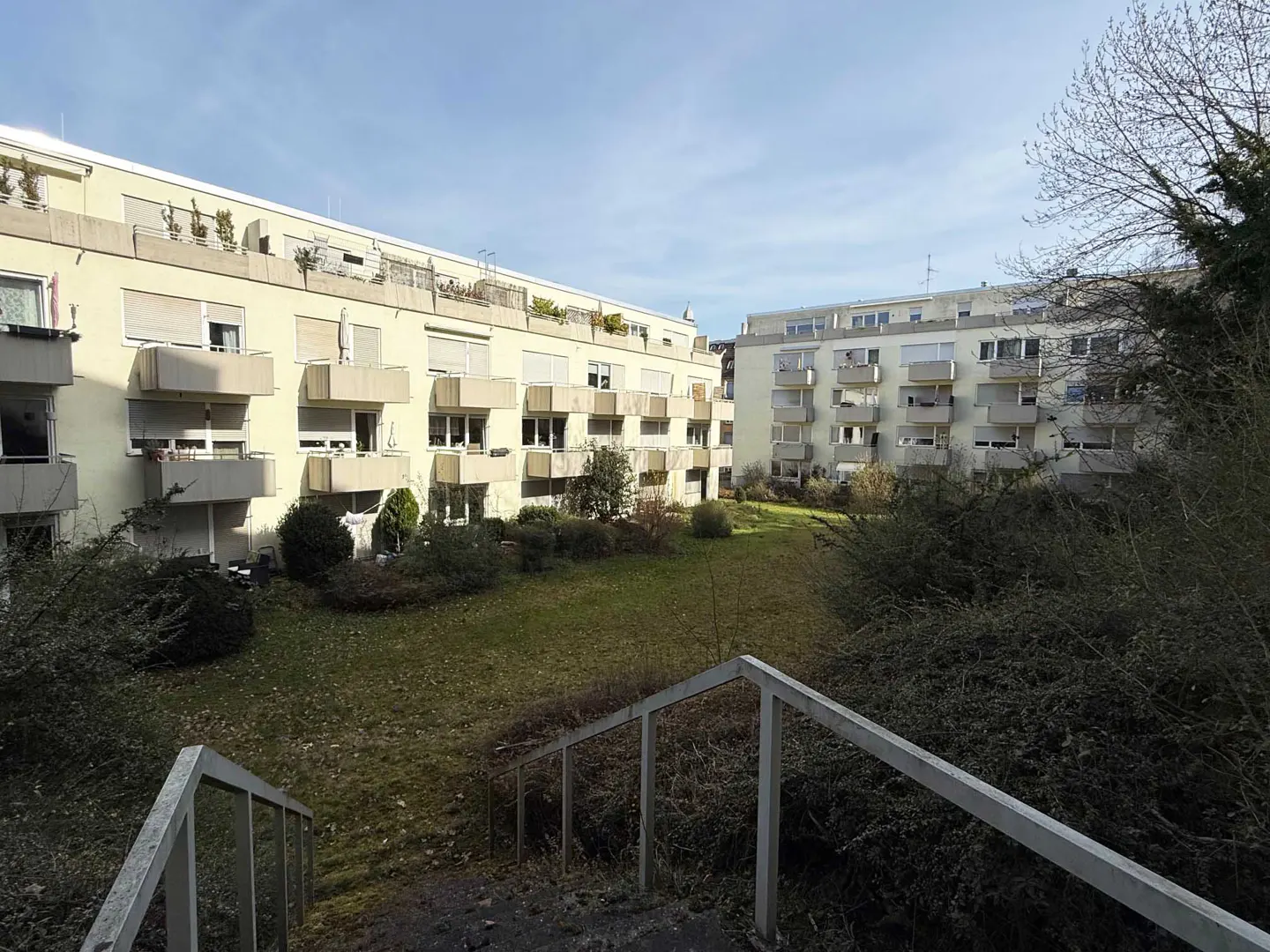 View of apartment buildings with balconies, green lawn, bushes, and metal stairs in the foreground.