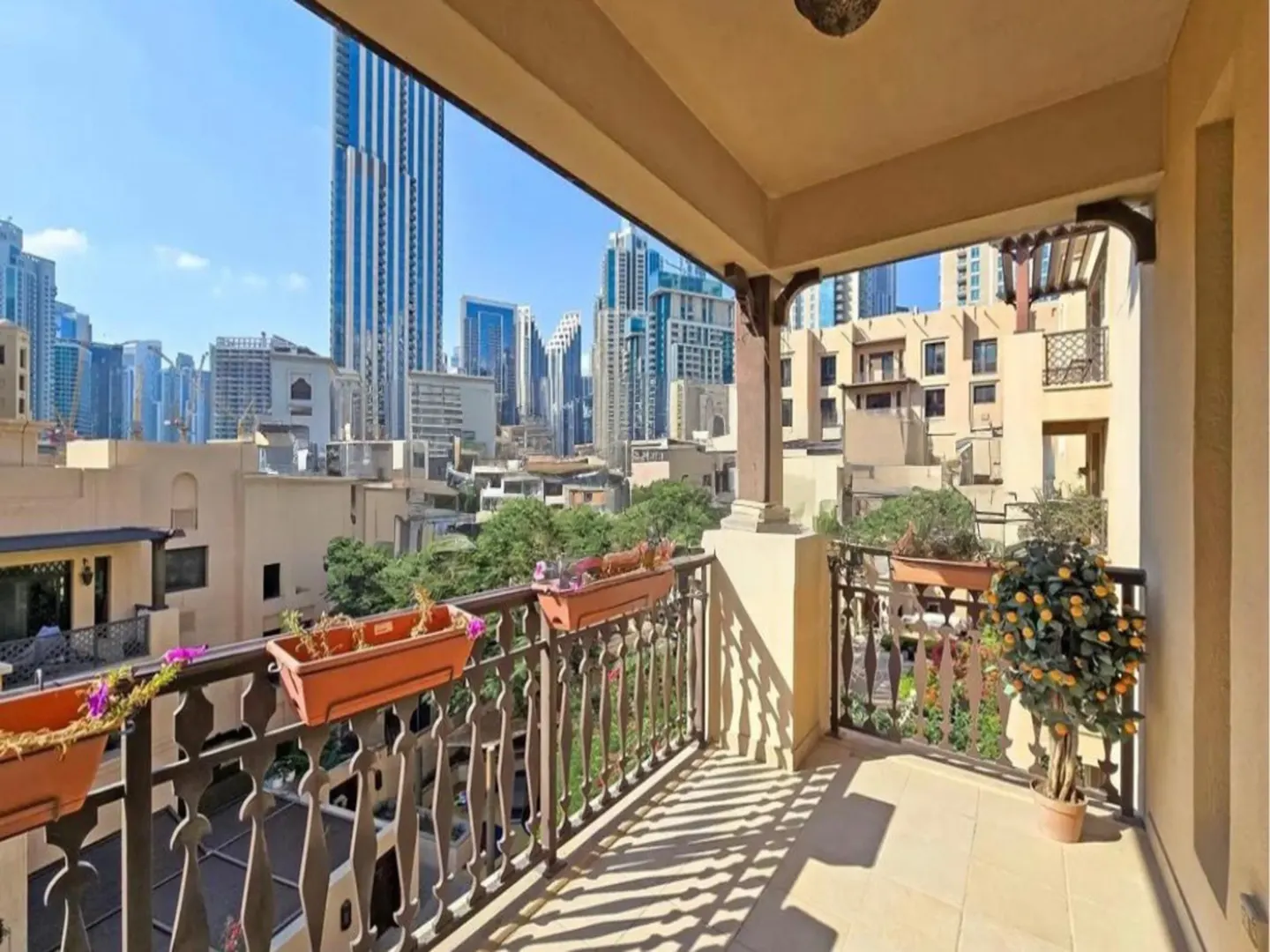 Balcony view of Dubai skyline. Ornate black railing with flower boxes. Beige tile floor and ceiling. Tall buildings in the background.