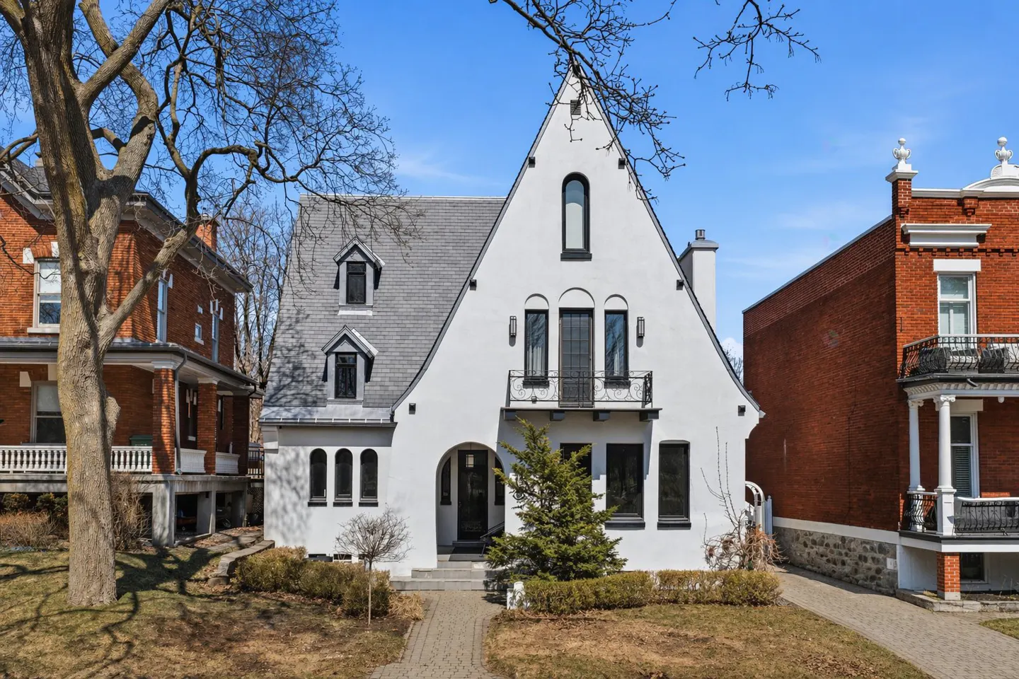 A white house with a gray roof and black trim stands between two brick houses on a sunny day.