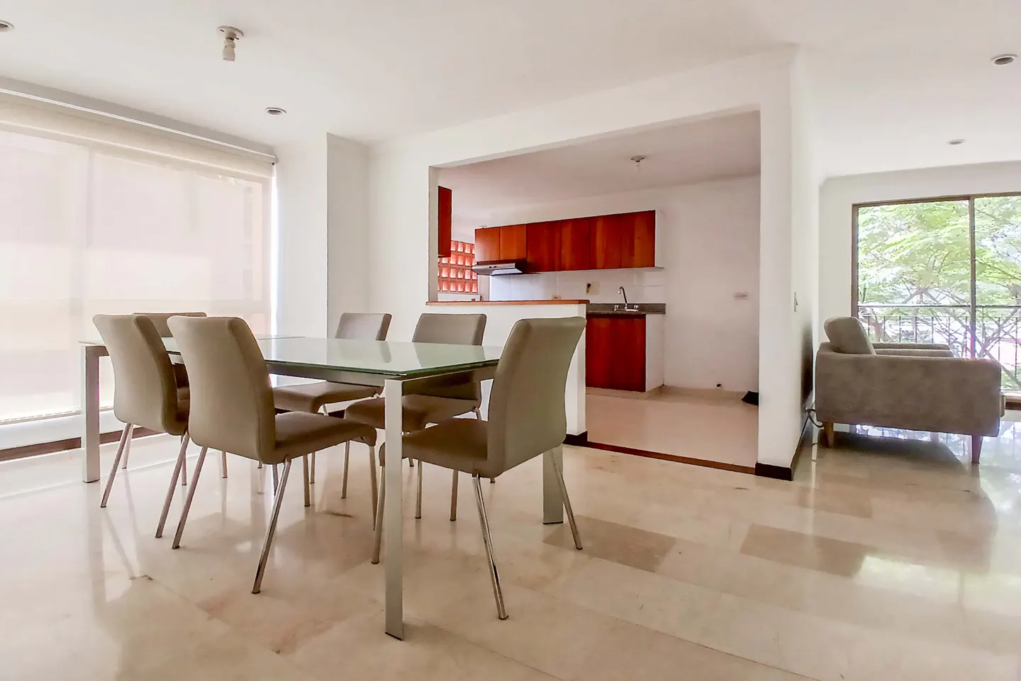 Bright, open-concept living space with a glass-top dining table, beige chairs, and a view into the kitchen with wood cabinets. A gray sofa sits near a window.