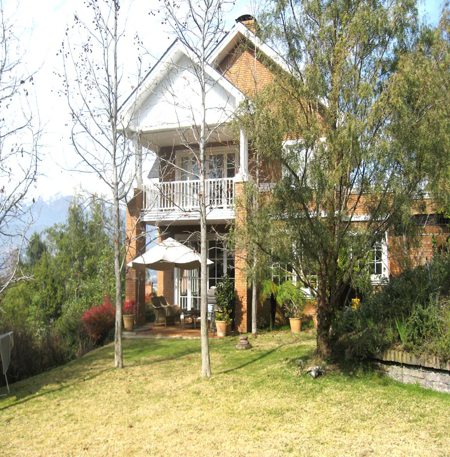 Two-story brick house with white trim, a balcony, and a patio with an umbrella, surrounded by trees and a lawn.