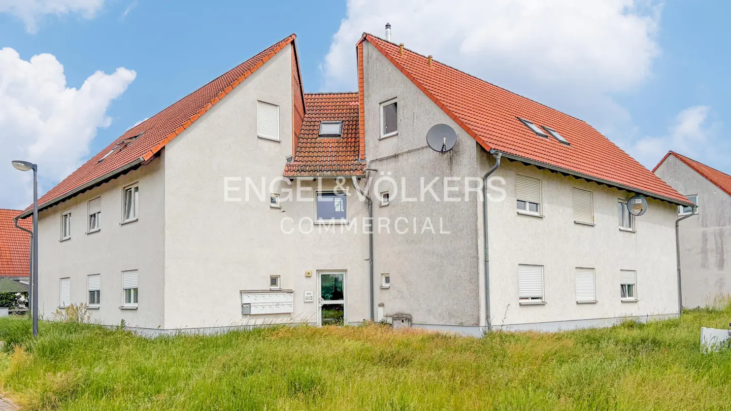 Two-story apartment building with white stucco walls, a red tile roof, and a grassy yard under a blue sky.