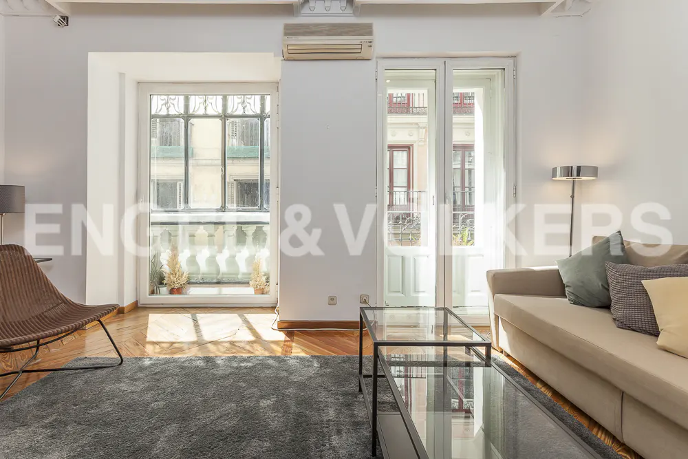 Bright living room with a gray rug, tan sofa, and glass coffee table. Sunlight streams through a window and balcony doors.