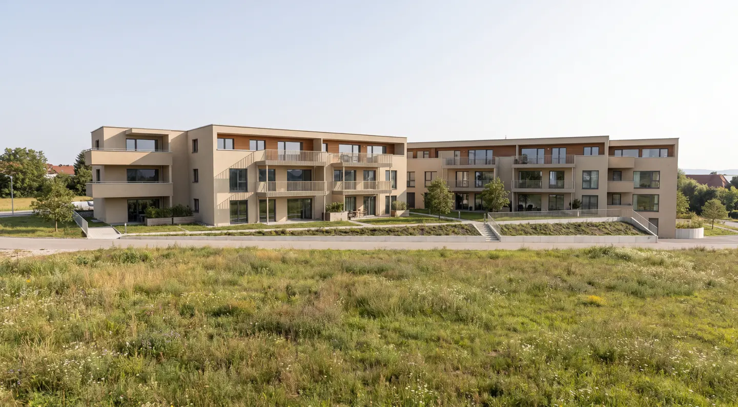 Modern beige apartment buildings with balconies and large windows, set behind a grassy meadow under a clear blue sky.